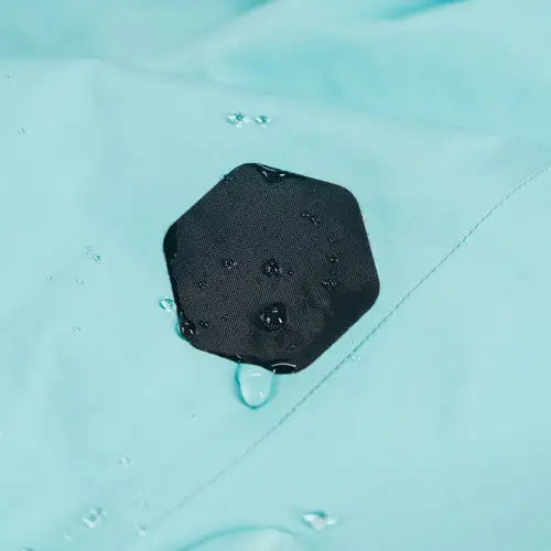 A black Gear Aid Tenacious Tape Hex Patch sticks to light blue fabric as water beads on and around it, highlighting Gear Aid’s ultra-aggressive adhesive and exceptional weatherproof tape.